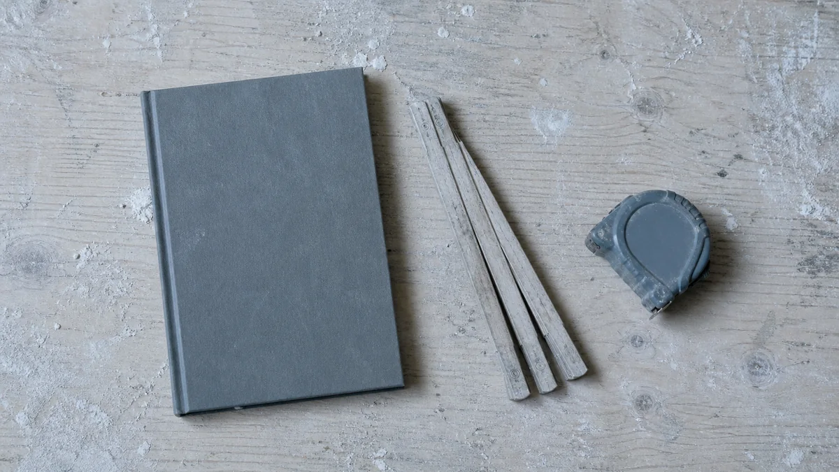 Cahier de chantier ouvert sur une table, couvert de notes manuscrites et de relevés de quantités, à côté d'un mètre et d'un crayon de charpentier
