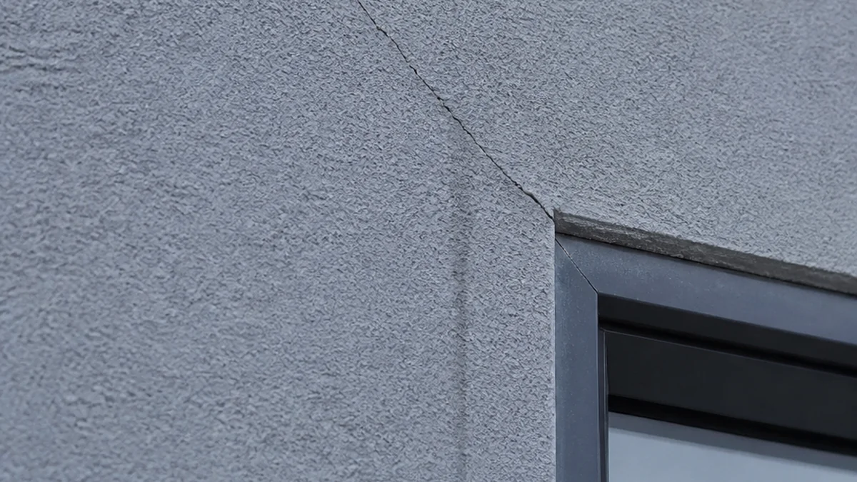 Fissure diagonale dans l'enduit extérieur fraîchement posé d'une maison neuve, près de l'angle d'une fenêtre, avec une légère trace d'humidité sombre en dessous suggérant un début d'infiltration