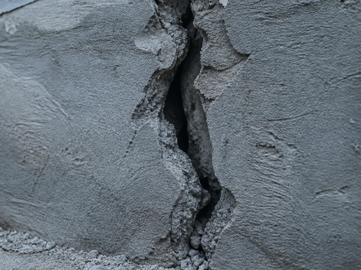 Fissure verticale profonde traversant toute l'épaisseur d'un mur en parpaing, gravats et poussière de ciment visibles au niveau de la brèche
