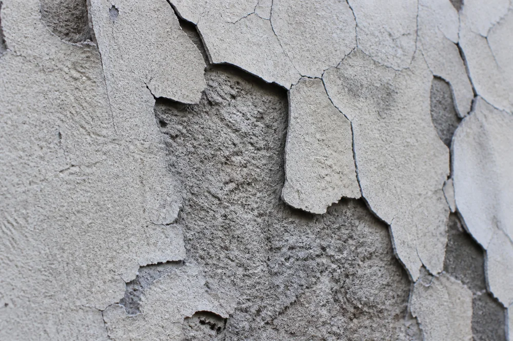 Gros plan macro sur un enduit de façade cloqué et décollé par plaques sur un mur crépi ancien, support apparent en dessous, légère fissuration en étoile