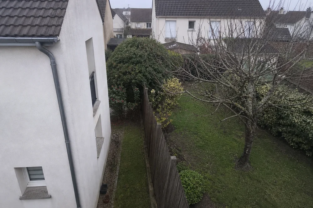 Vue en surplomb d'un jardin résidentiel montrant l'angle d'une maison construite près de la limite séparative avec la parcelle voisine, toits de maisons voisines en arrière-plan, aucune personne visible