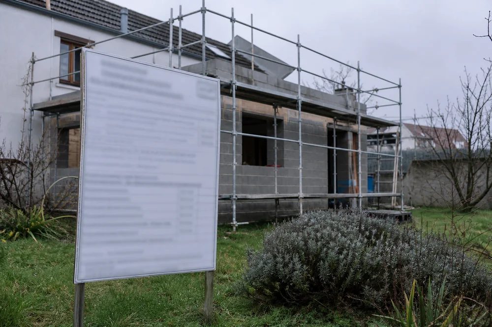 Panneau d'affichage réglementaire de permis de construire planté dans un jardin devant une extension de maison en construction, échafaudage en arrière-plan, aucune personne visible